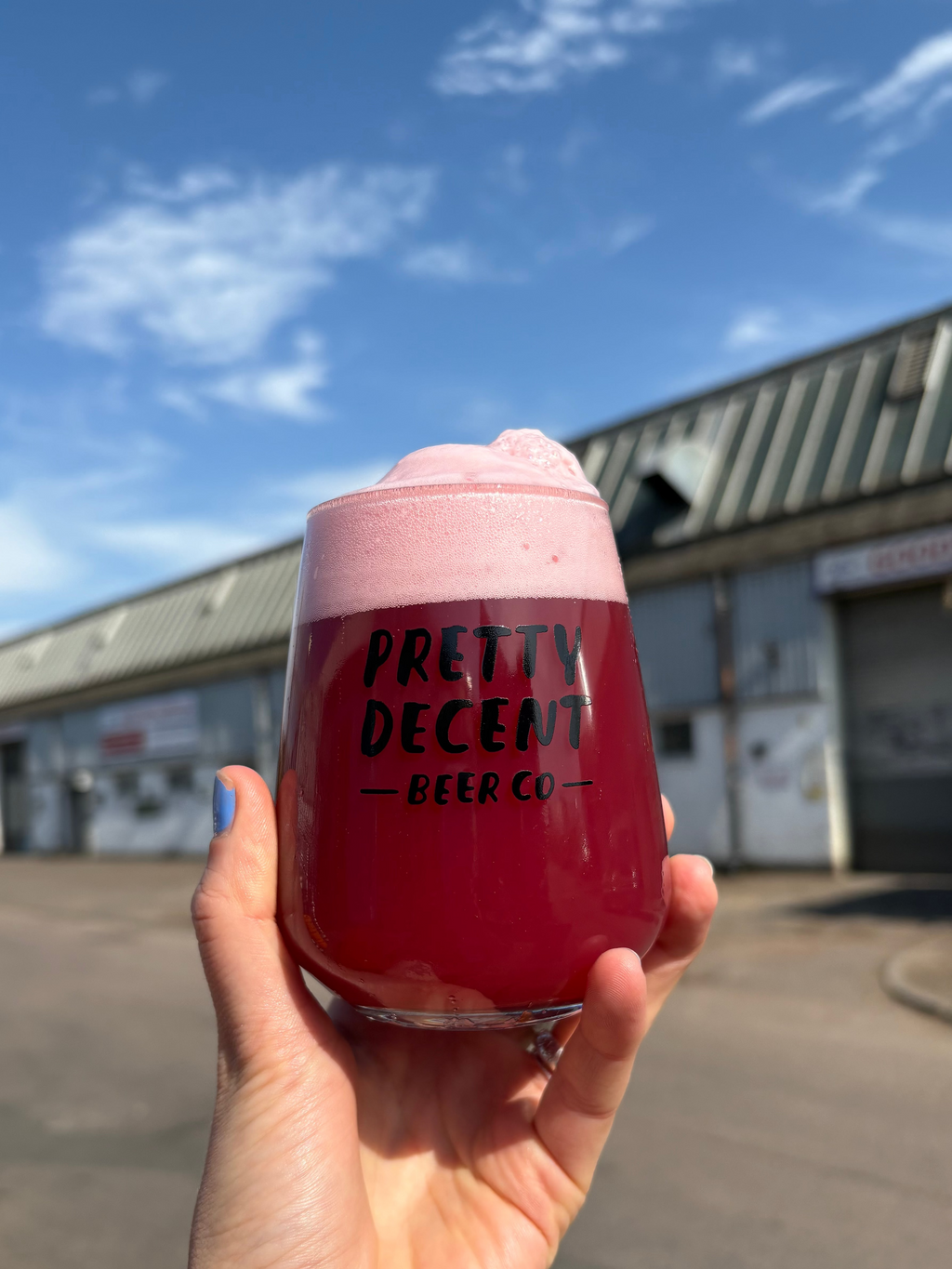 Hand holding a pink drink in a cup with 'Pretty Decent Beer Co.' text against a warehouse background.